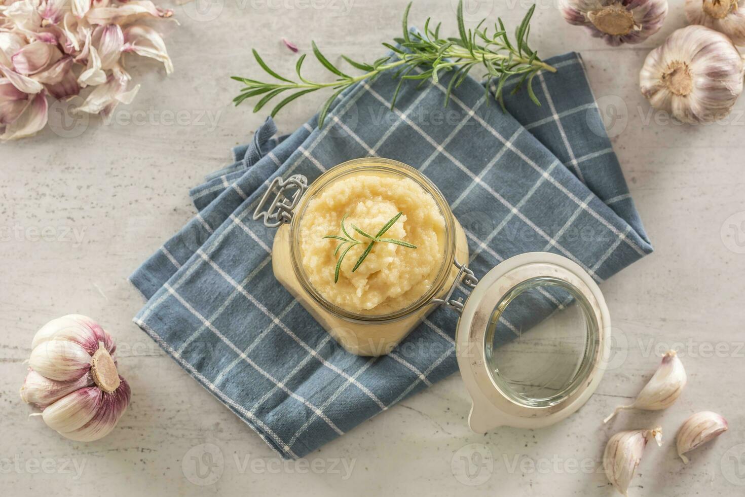 Top view aromatic garlic paste in a glass jar laid on rustic kitchen cloth with bulbs and peeled cloves and rosemary photo