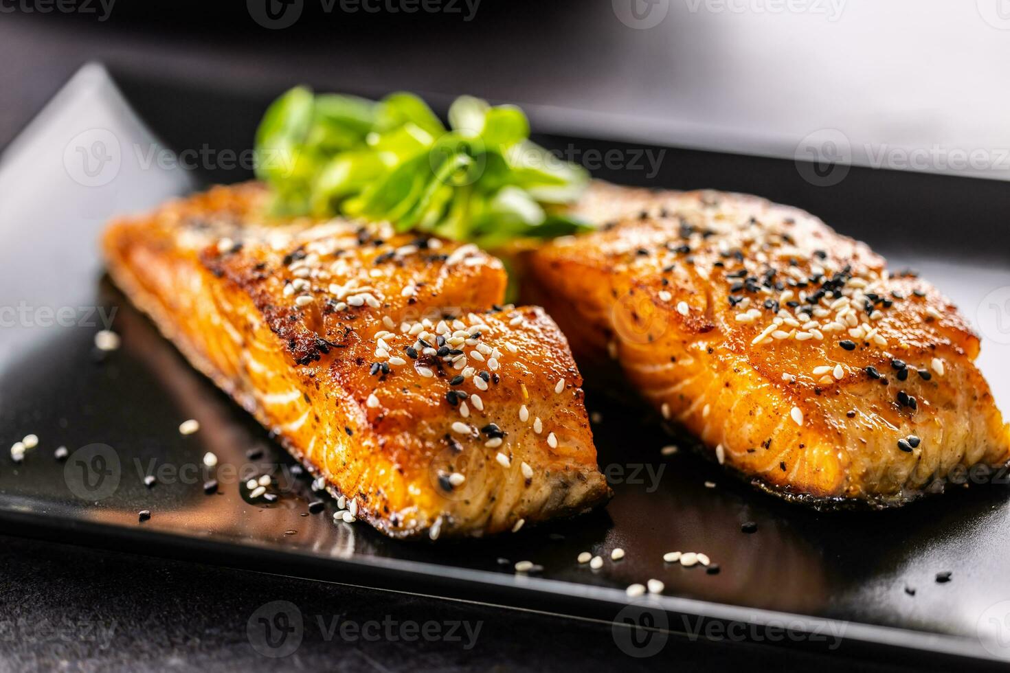 Two salmon fillets baked until crispy, sprinkled with sesame on a black plate. photo