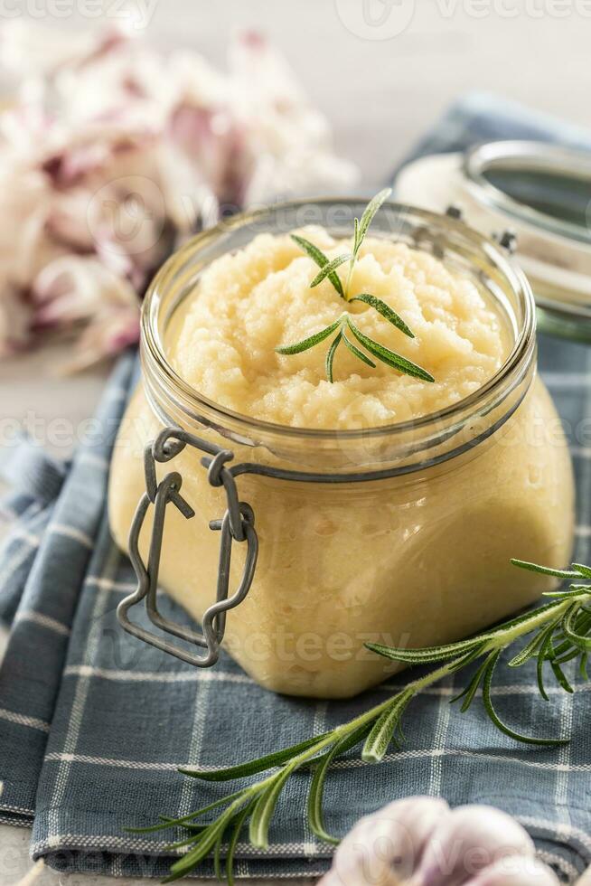 Aromatic garlic paste in a glass jar laid on rustic kitchen cloth with bulbs and peeled cloves and rosemary photo