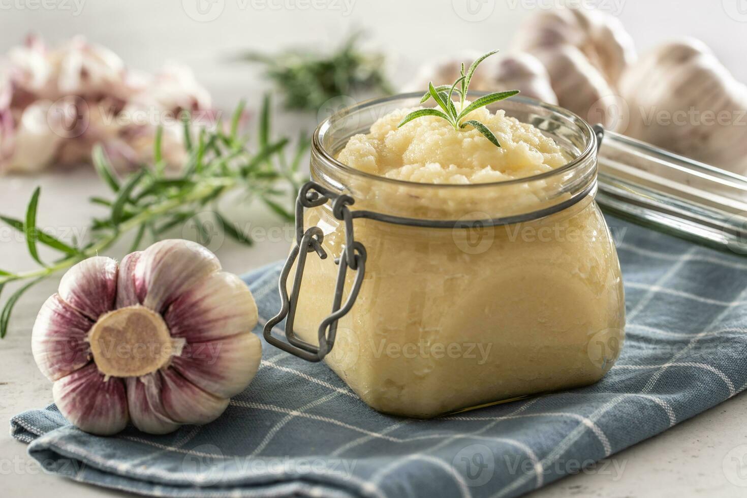 Aromatic garlic paste in a glass jar laid on rustic kitchen cloth with bulbs and peeled cloves and rosemary photo