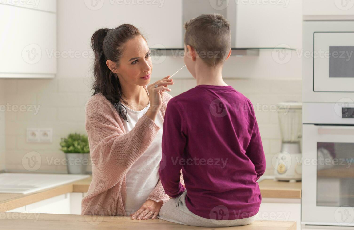 Home testing for Covid-19 as mum puts swab into the nose of her son sitting on a kitchen desk photo