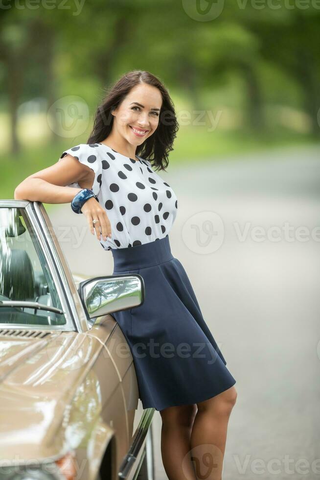 Good-looking brunette leaning her back and arm against a vintage cabriolet seductively looking into the camera photo