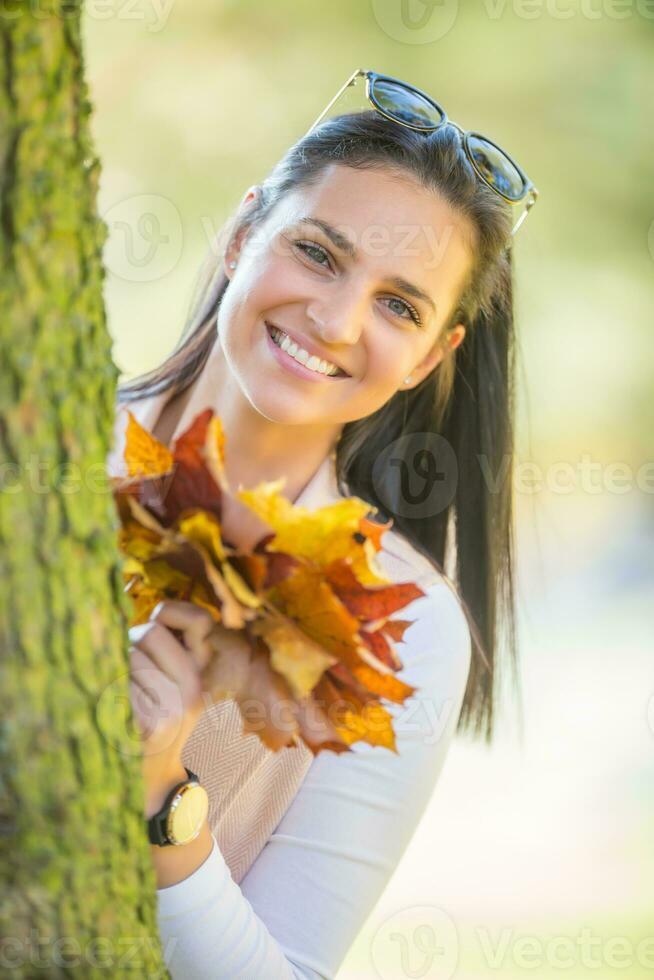 Happiness woman sitting in autumn park holding bouquet with fall leaves cute look from behind a tree photo