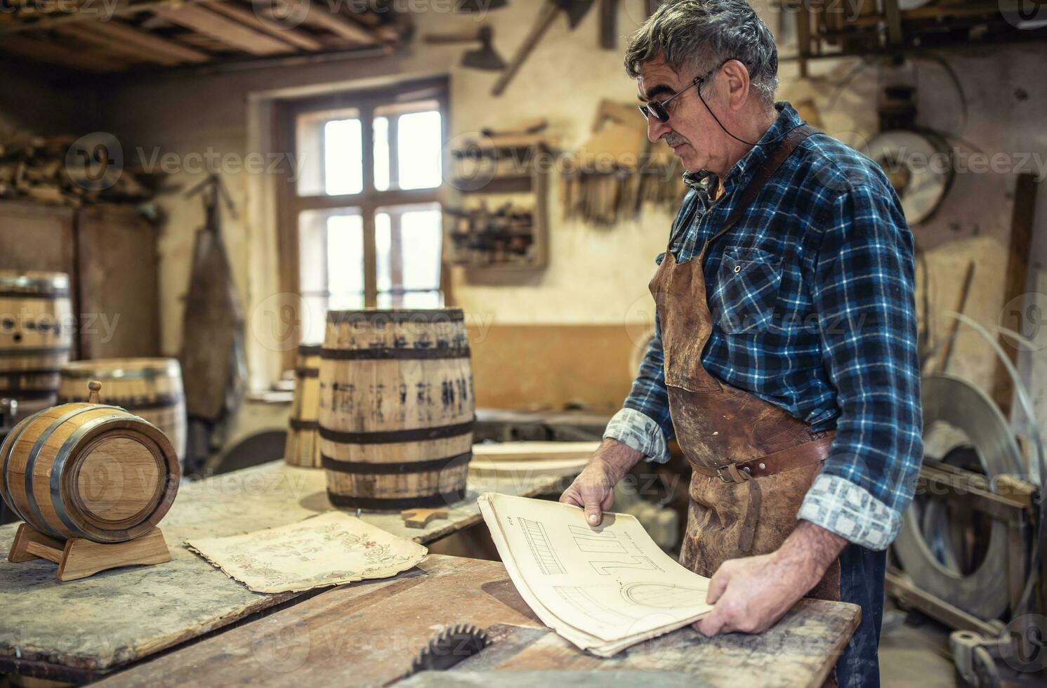 Aged craftsman reads drawings in his rustic wooden barrels producing workshop photo