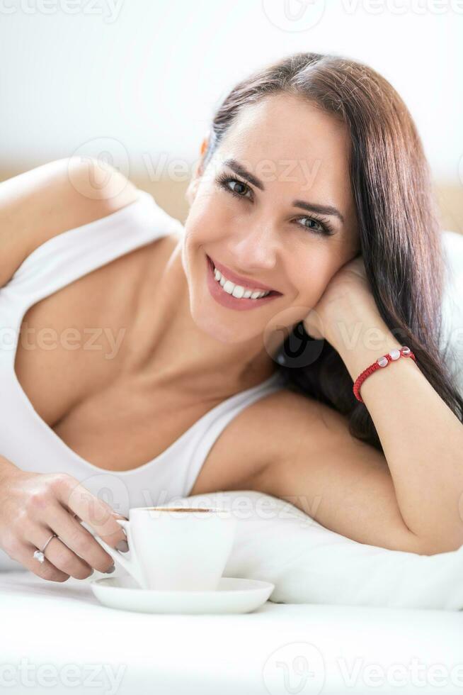 hermosa morena en blanco camisa mentiras en su lado teniendo un taza de café, sonriente foto