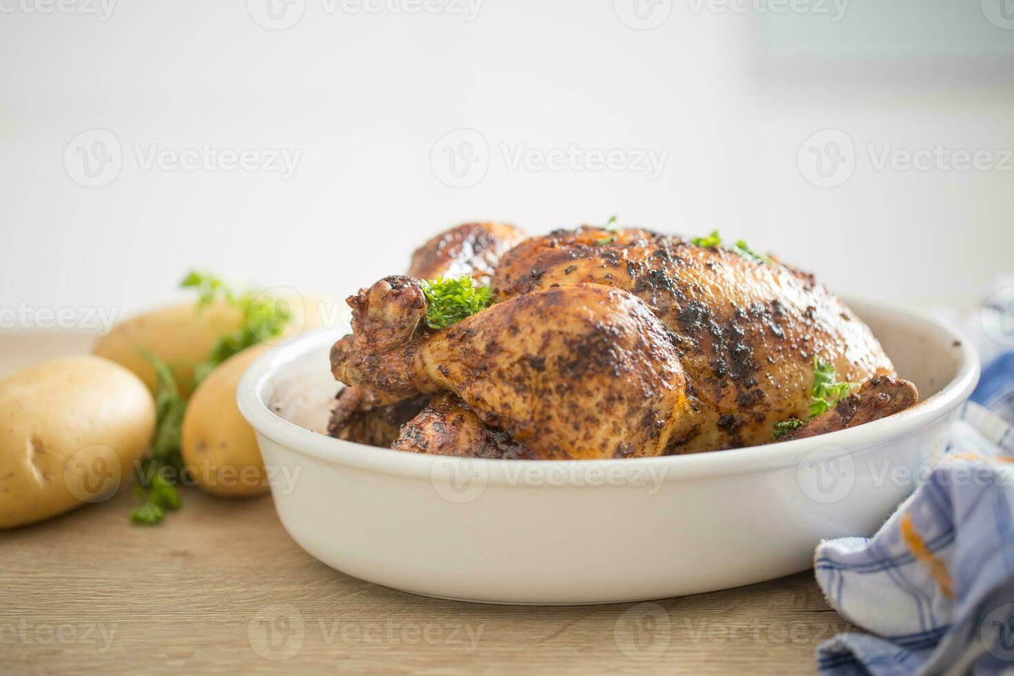 Whole chicken roasted in white dish with raw potatoes on kitched table. Home interior in the background photo