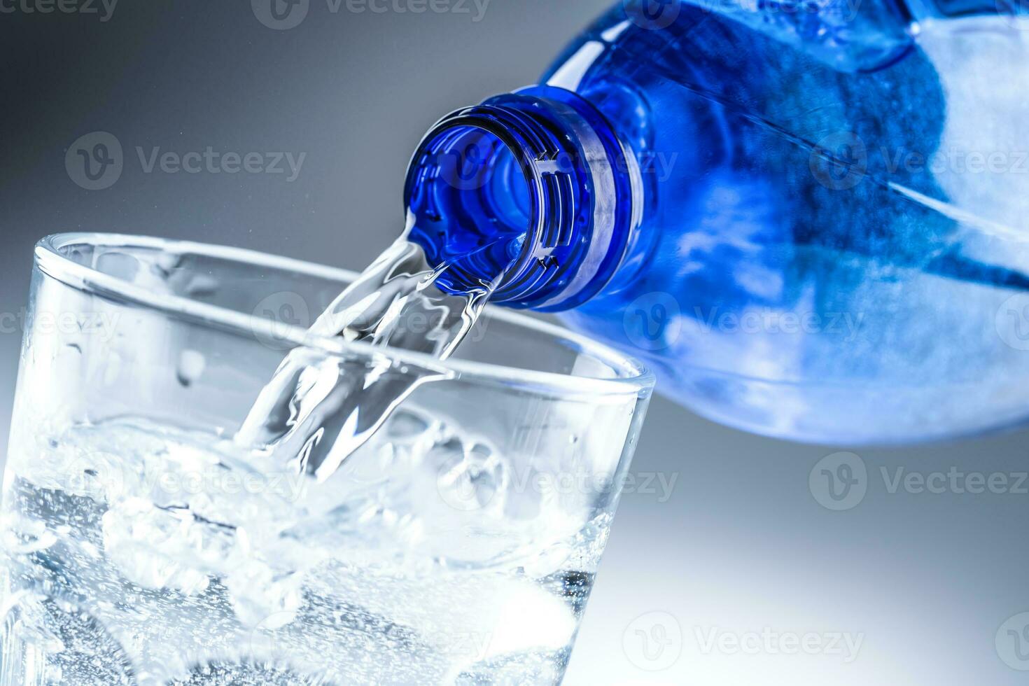 Pouring mineral water from blue bottle into clear glass on abstract grey background photo