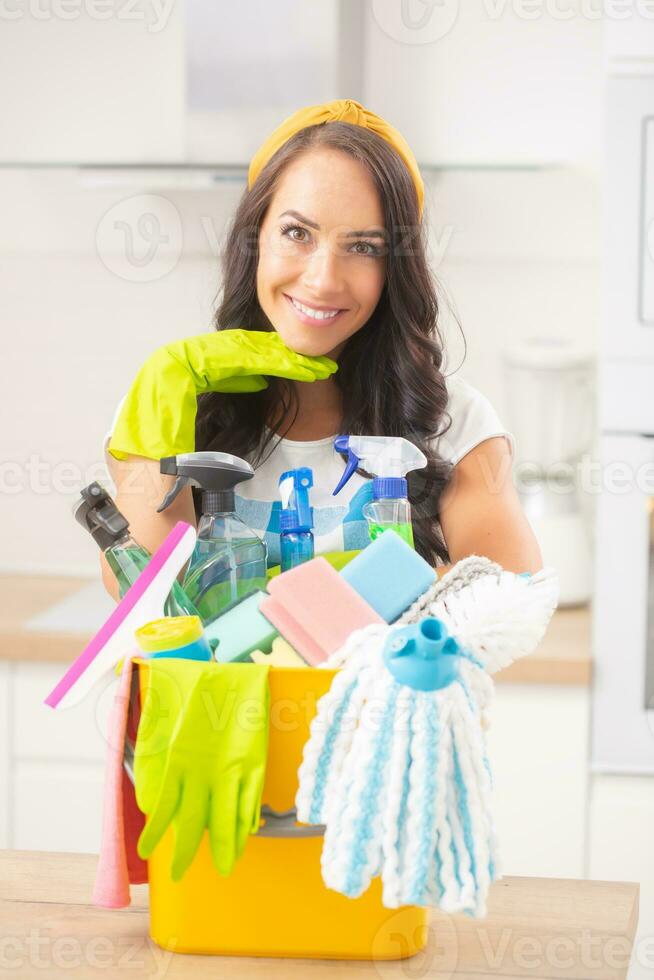 Portrait of a good-looking female with head resting on a rubber glove, smiling and leaning against a bucketful of cleaning stuff photo