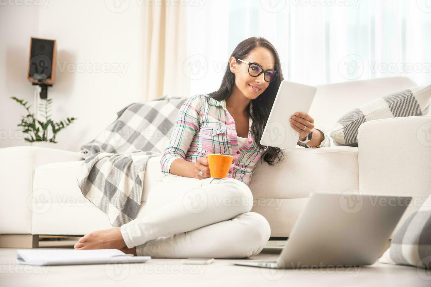 Good looking girl in glasses reading a tablet and drinking coffee leaned against a couch and sitting on a floor photo