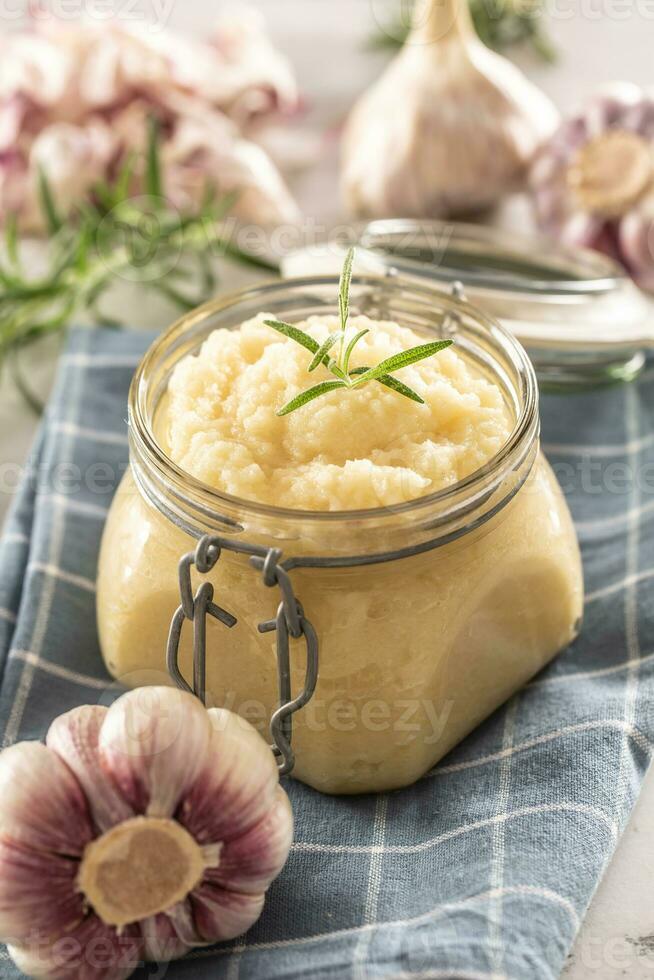 Aromatic garlic paste in a glass jar laid on rustic kitchen cloth with bulbs and peeled cloves and rosemary photo