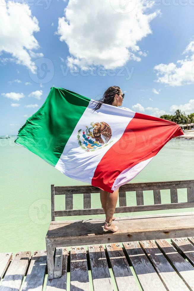 Woman holds Mexican flag behind her back as she stands on a bench on a pier next to the Carribean Sea photo