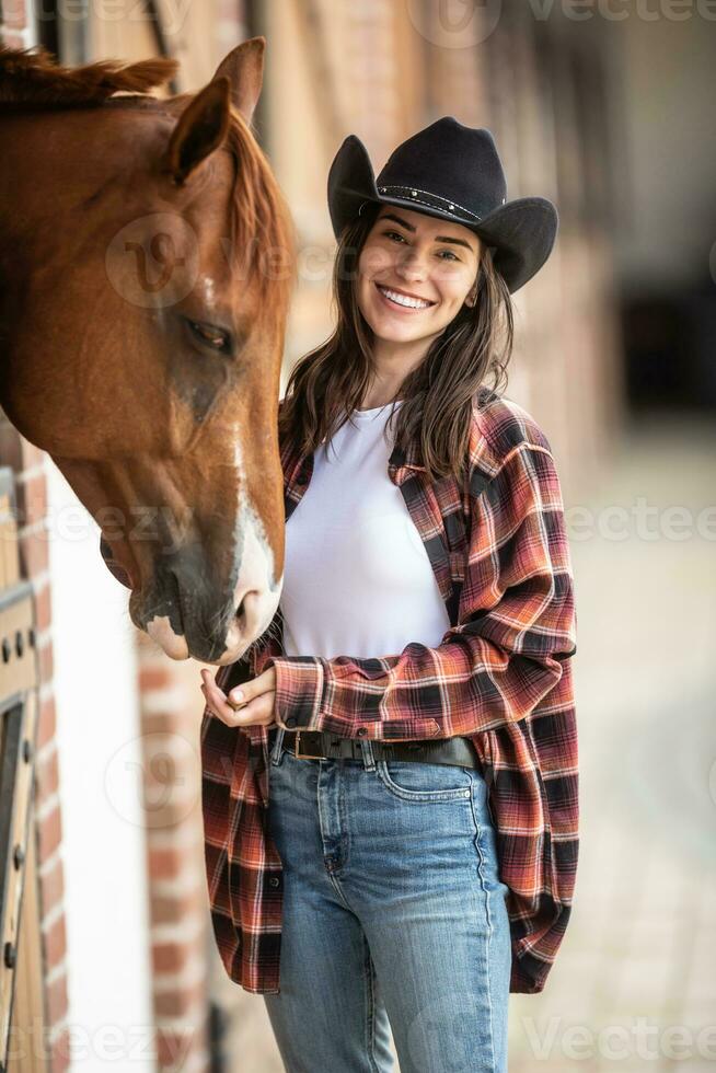 vaquera con un grande sonrisa alimenta su caballo desde el mano dentro el estable foto
