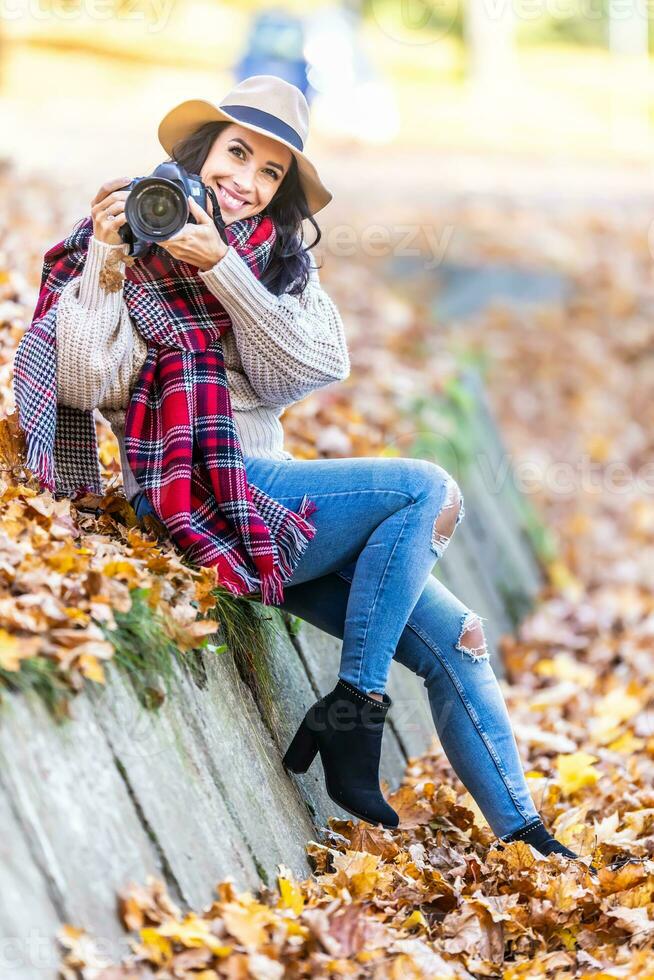 Smiling good-looking female photographer in a hat and stylish autumn clothes takes pictures outside on a sunny fall day photo