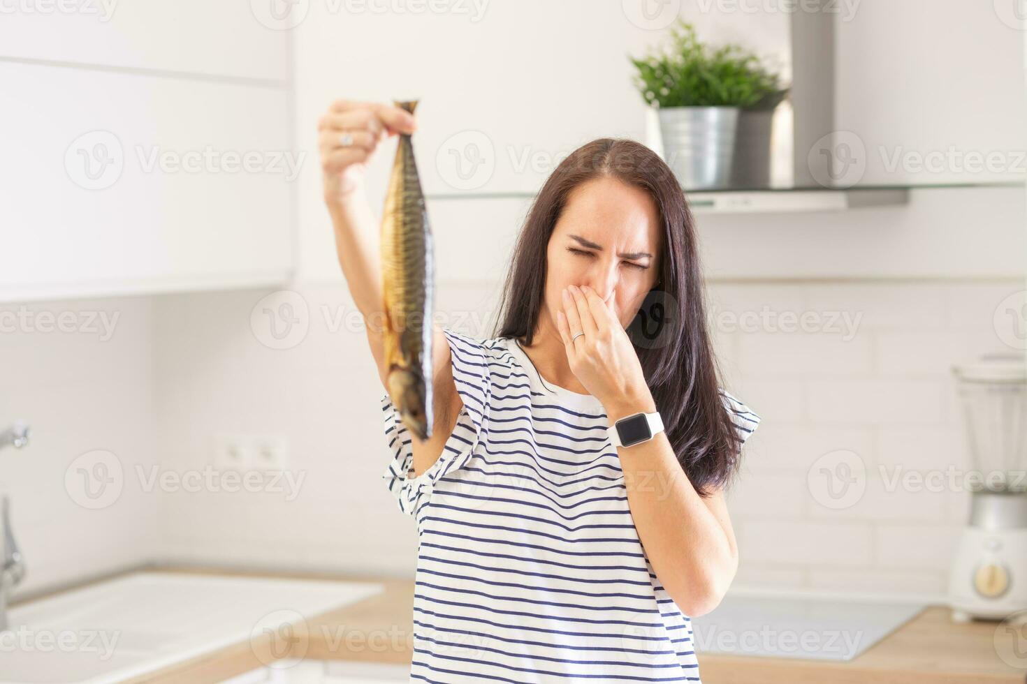 Woman disgusted from a smell of a fish she holds in hand holds her nose standing in the kitchen at home photo