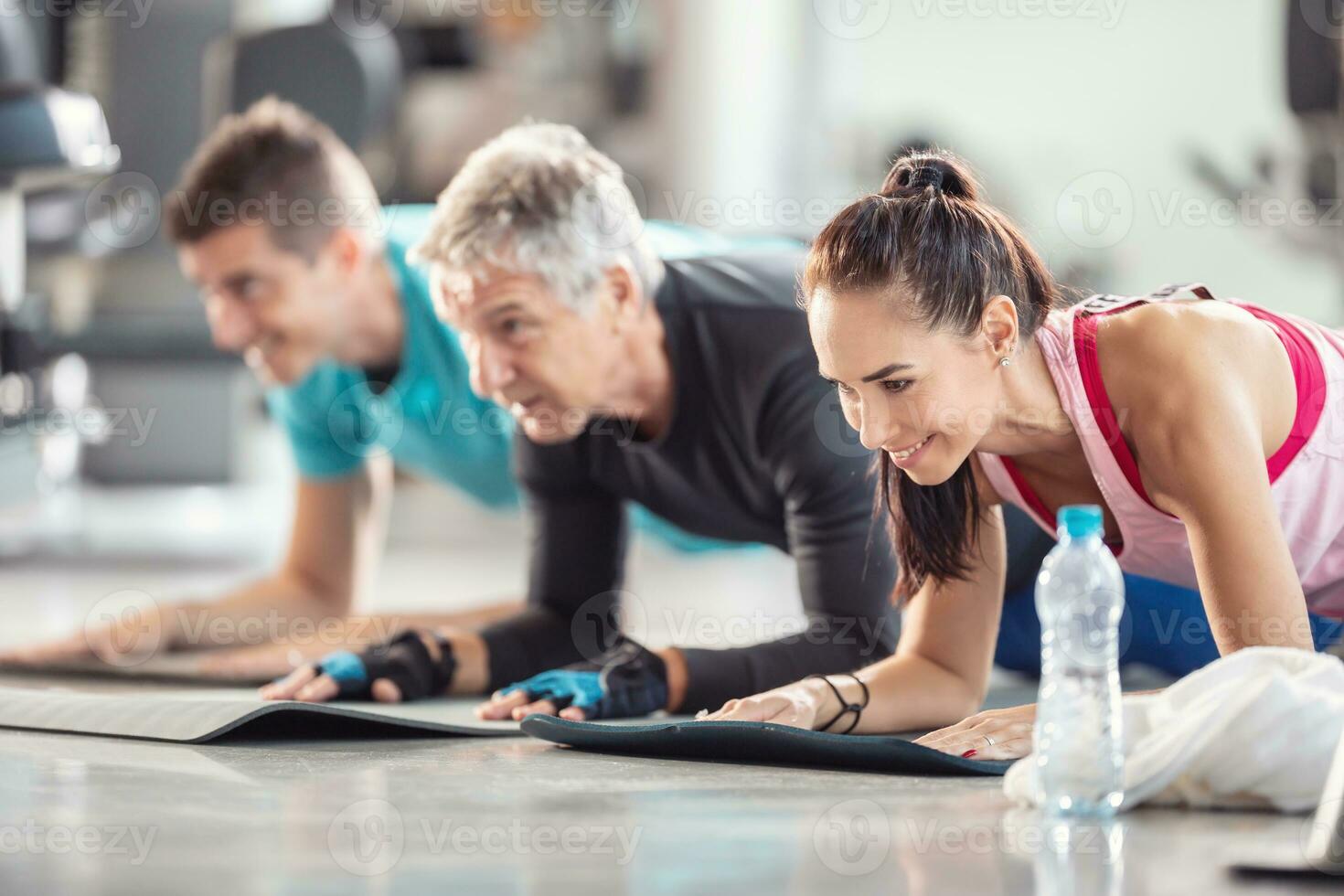 Diverse age group doing elbow planks on mats inside the gym with a plastic water bottle in the front photo