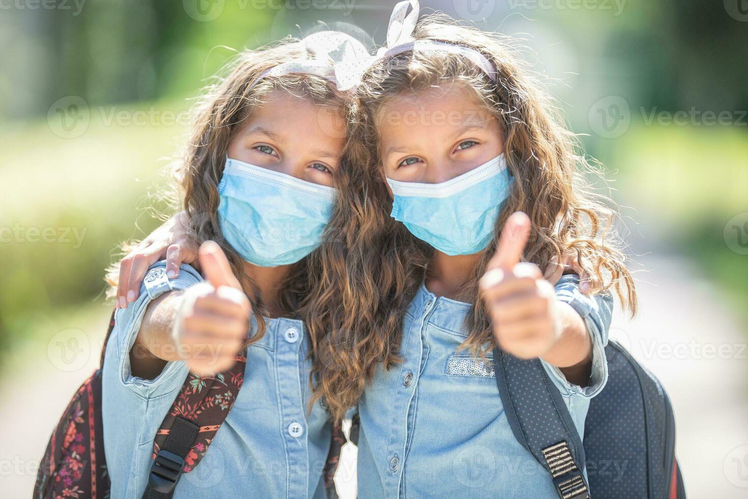 Twin sisters with face masks go back to school during the Covid-19 quarantine and showing thumbs up photo