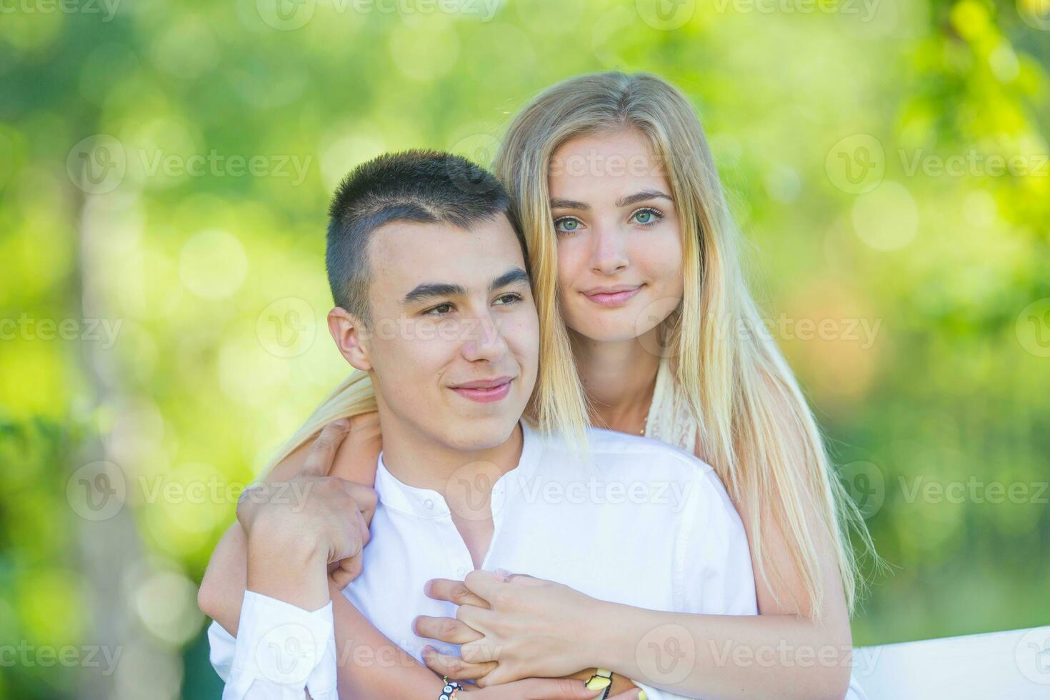 Beautiful young girl hugging her dark-haired boyfriend from behind while holding hands in the nature. Girl looking into the camera while boy is looking to the side photo