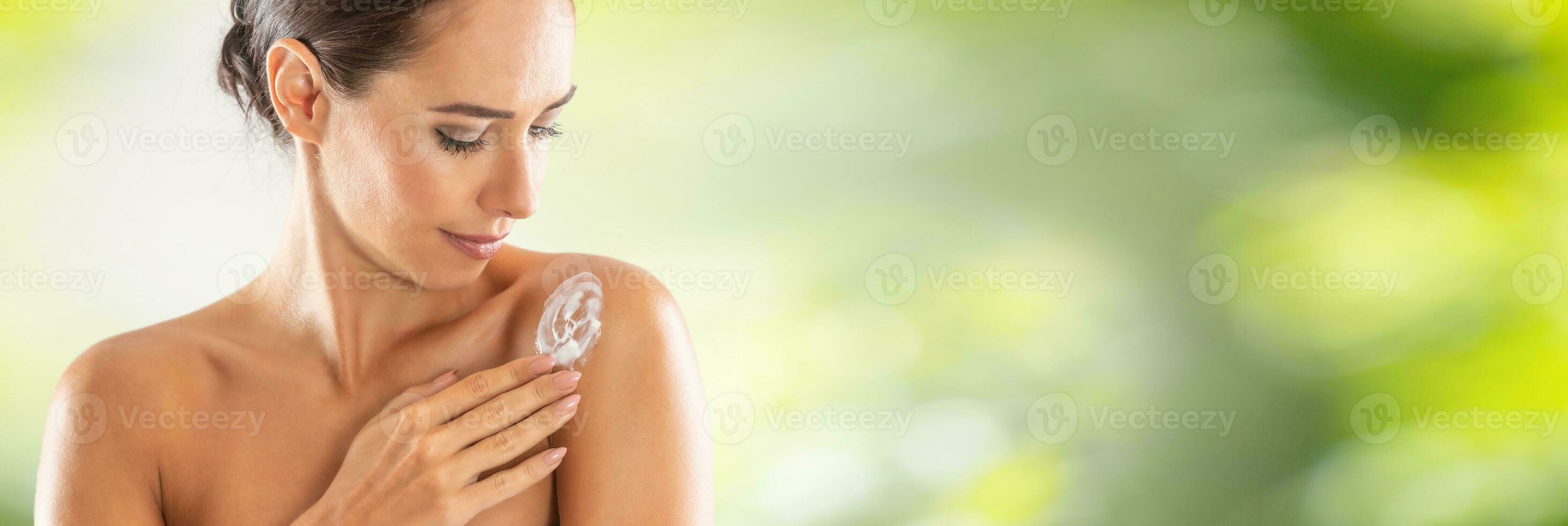 Beautiful young woman applying cream to her shoulder. Skincare spa wellness and cosmetics concept. Blurred green panoramic banner photo