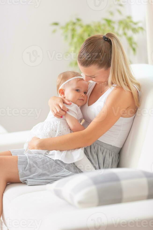 Young female blonde mom holds her baby daughter in her arms sitting on a sofa at home photo