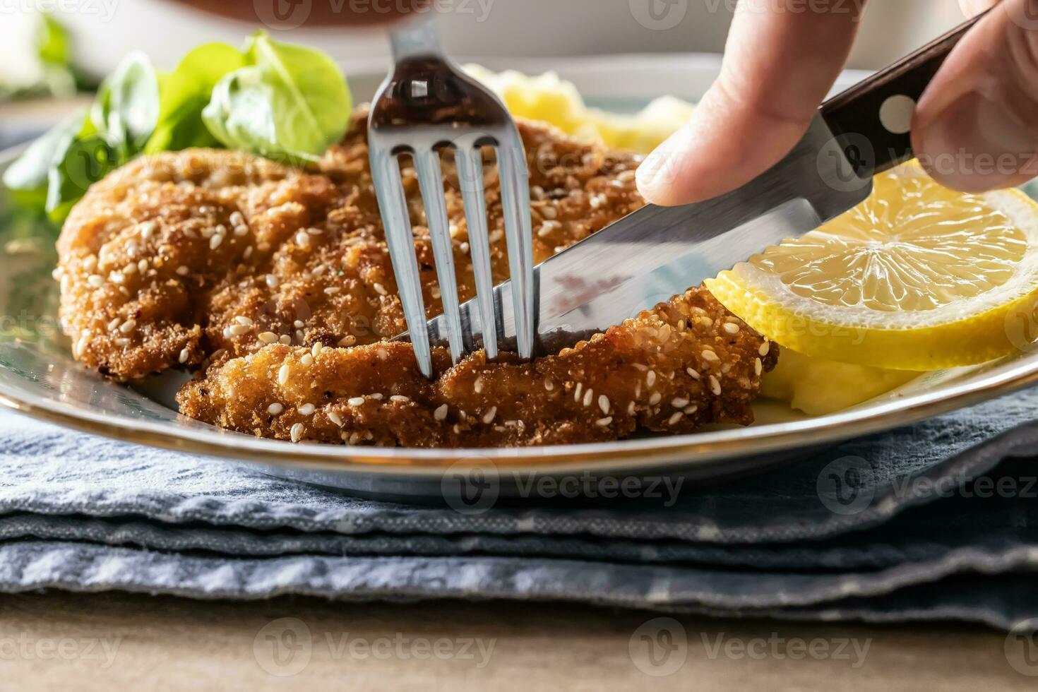 Fork and knife taking a first cut from a schnitzel in sesame seeds served with a slice of lemon photo