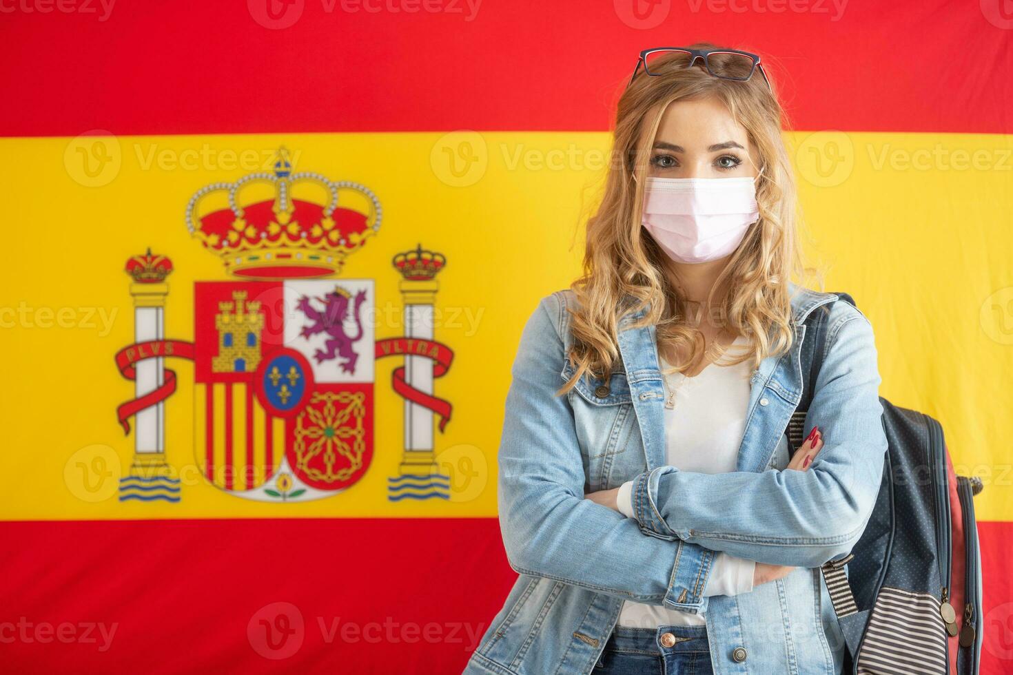 Female student caryying backpack stands in front of the Spanish flag with a face mask on photo