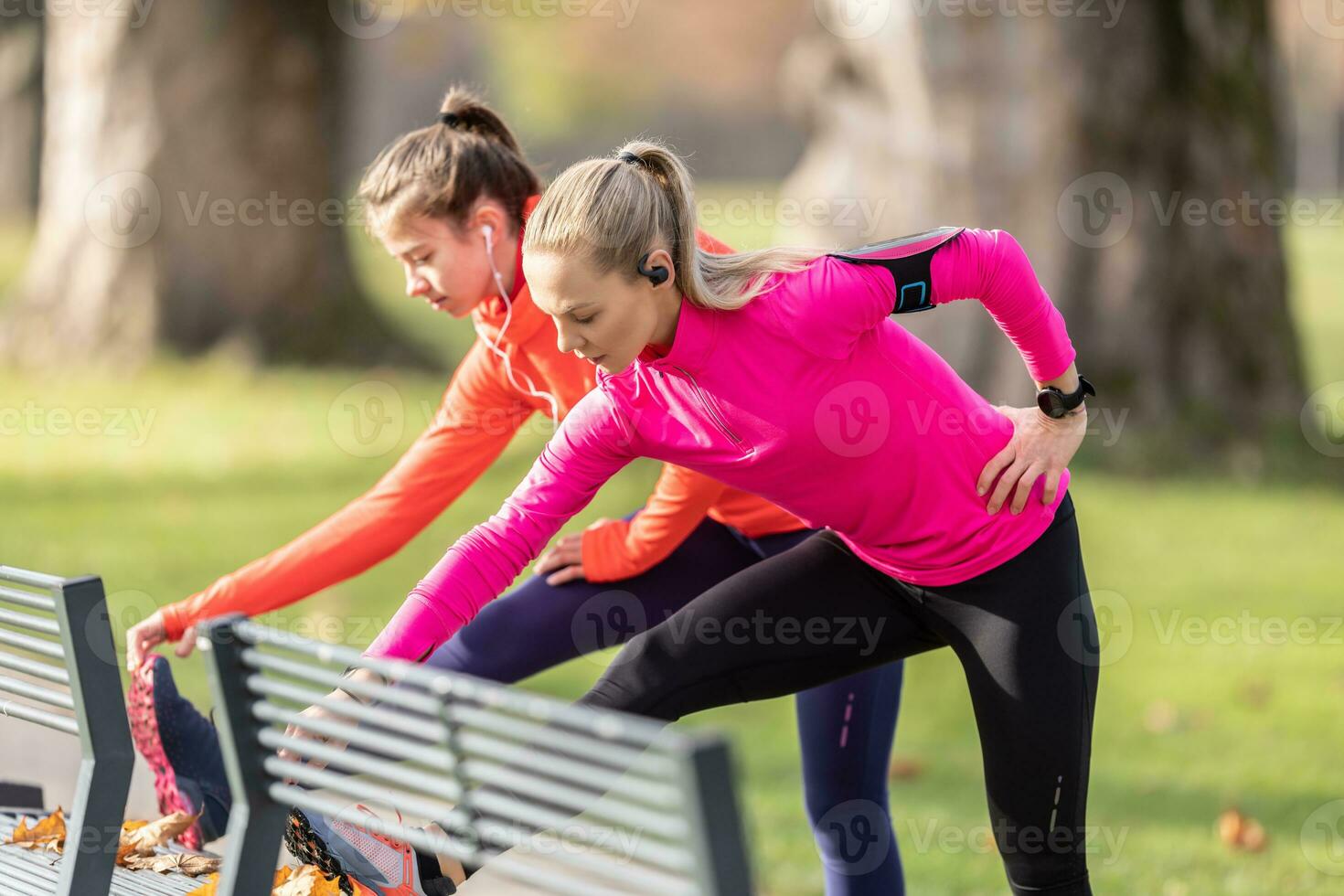 Two female athletes are warming up in the autumn park, using a bench to warm up before running photo