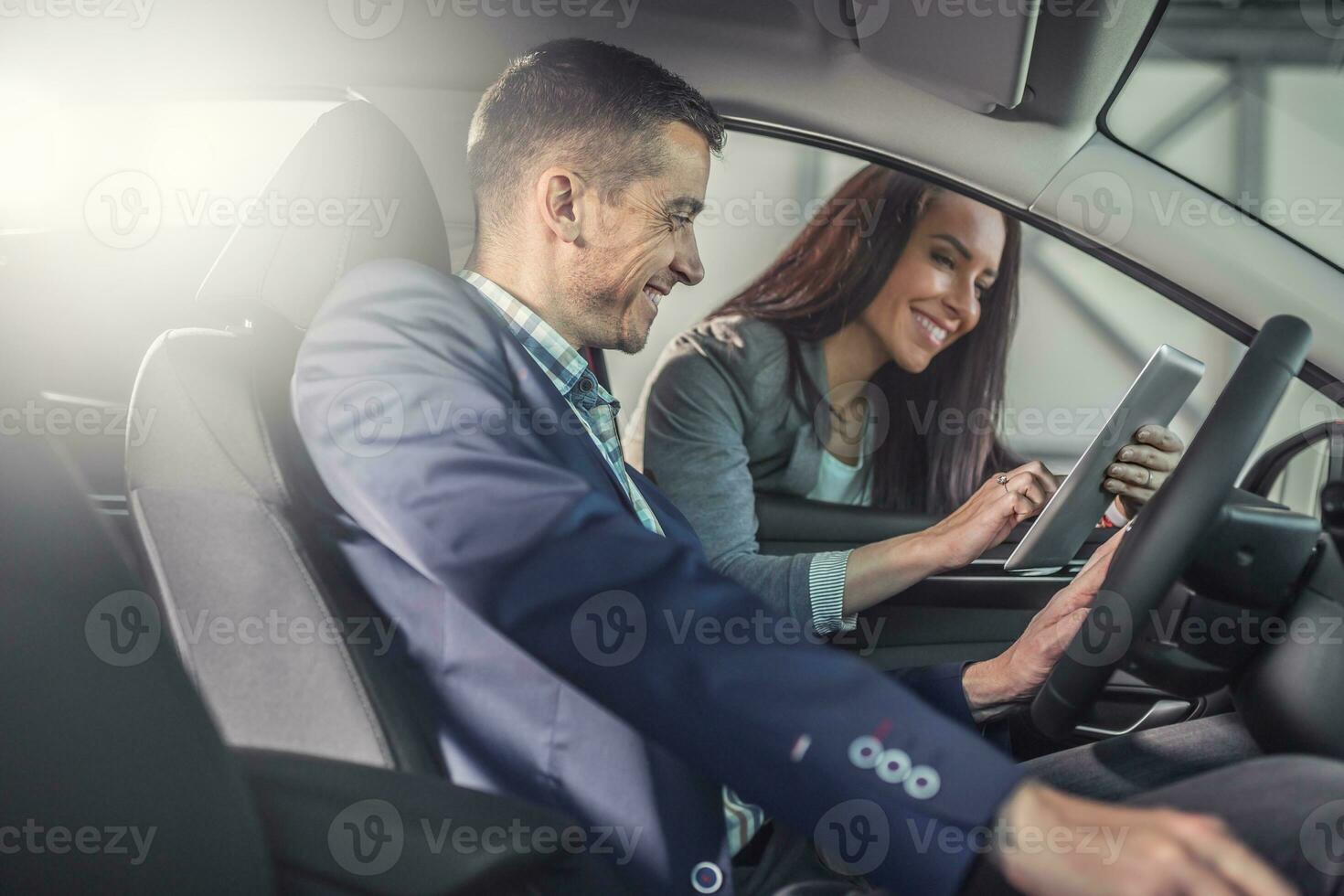 Man testing a car in the sales shop smiles looking into a tablet in the hands of the female car dealer showing technicalities photo