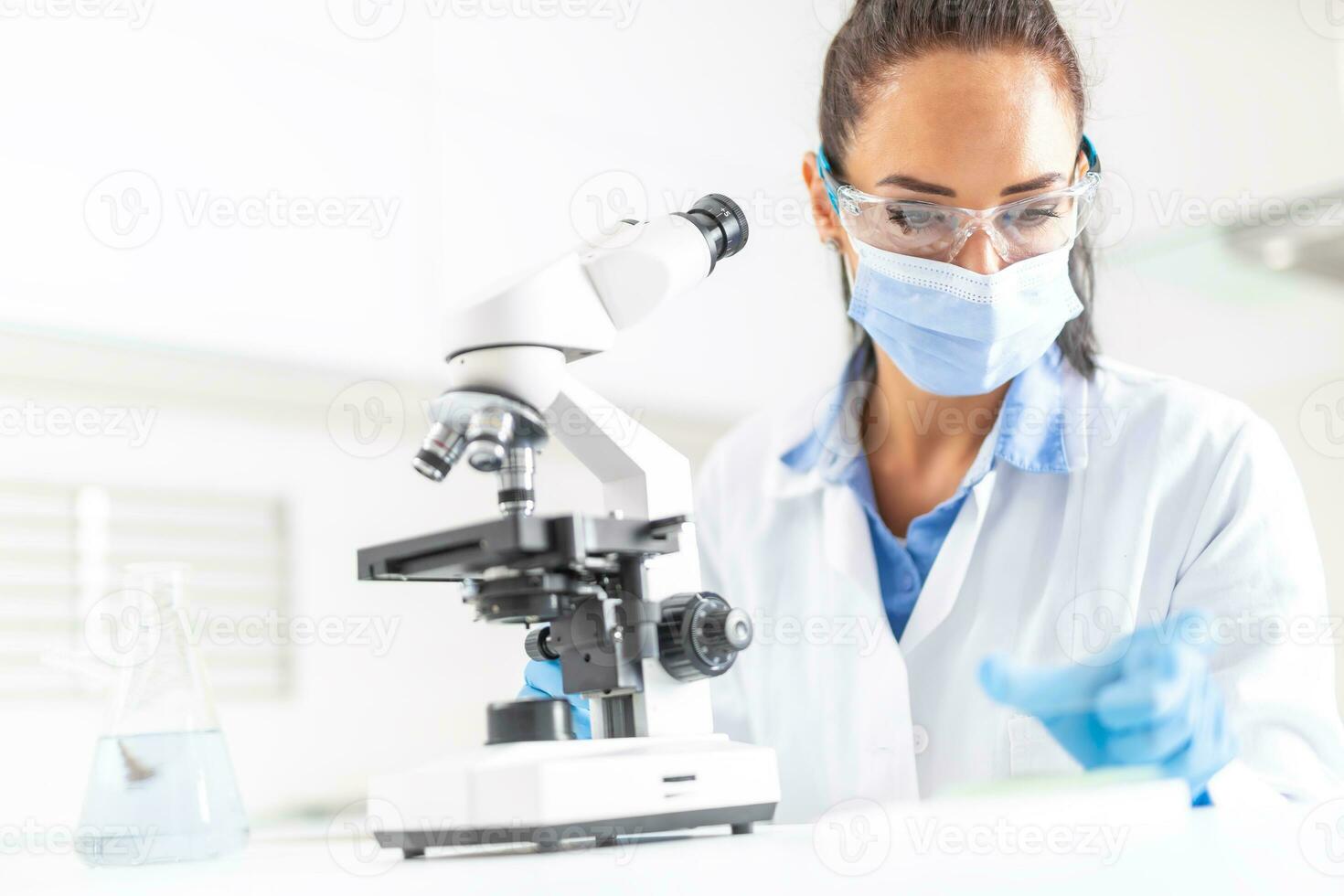 Female lab technician in protective glasses, gloves and face mask sits next to a microscope and conical flask, looking aside on the desk photo