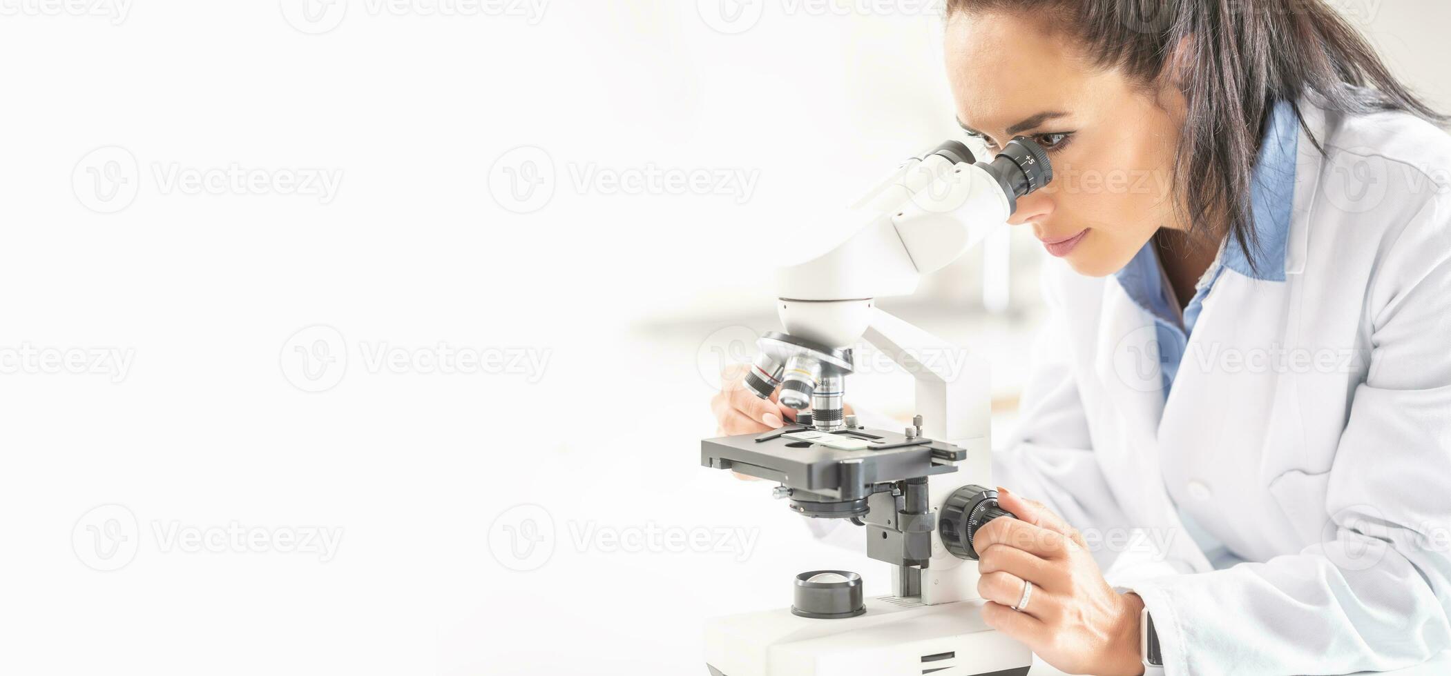 A female laboratory researcher looks into a microscope covering right half of the picture photo