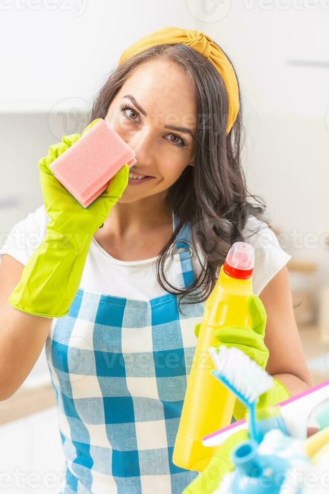 Sexy young woman smiling into the camera, holding a dish washing sponge in her rubber gloves, partially covering her face. Holding detergent in the other hand with more cleaning stuff in front of her photo
