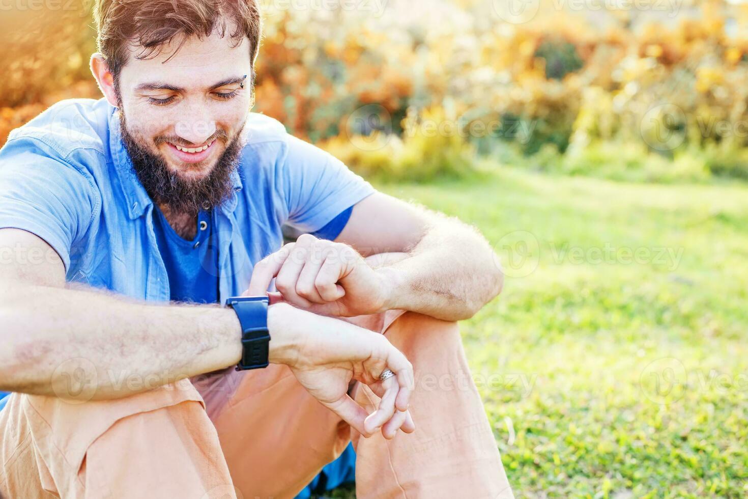 a man with a beard sitting on the grass looking at his watch photo