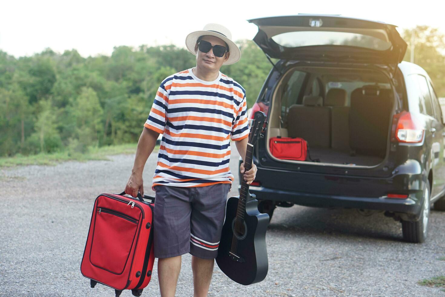 Asian man traveler goes traveling by car, holds red suitcase and the guitar. Concept, relax time. Vacation, holiday or weekend trip by private vehicle. Transportation. Road trip.Give time for yourself photo