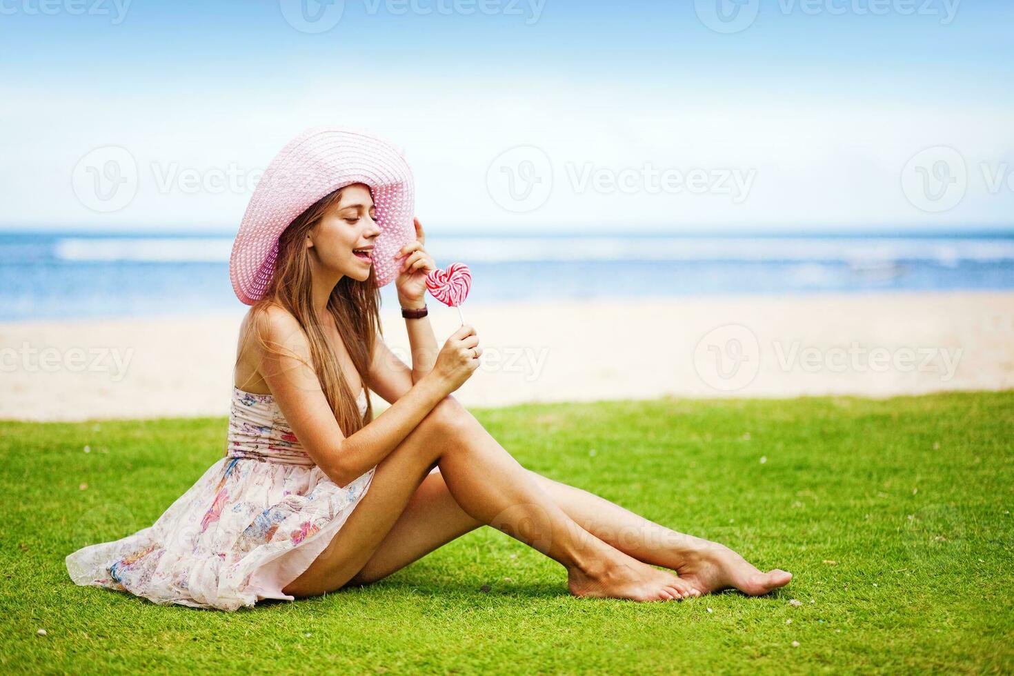 a woman in a pink hat sitting on the grass eating a lollipop photo