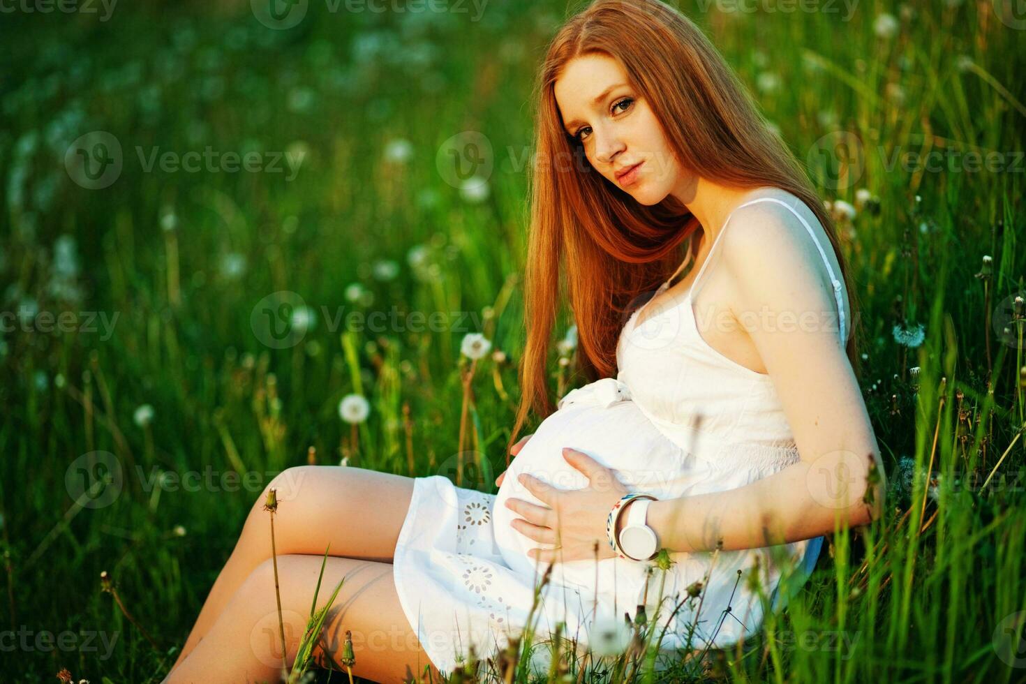 un embarazada mujer sentado en un campo con diente de león foto