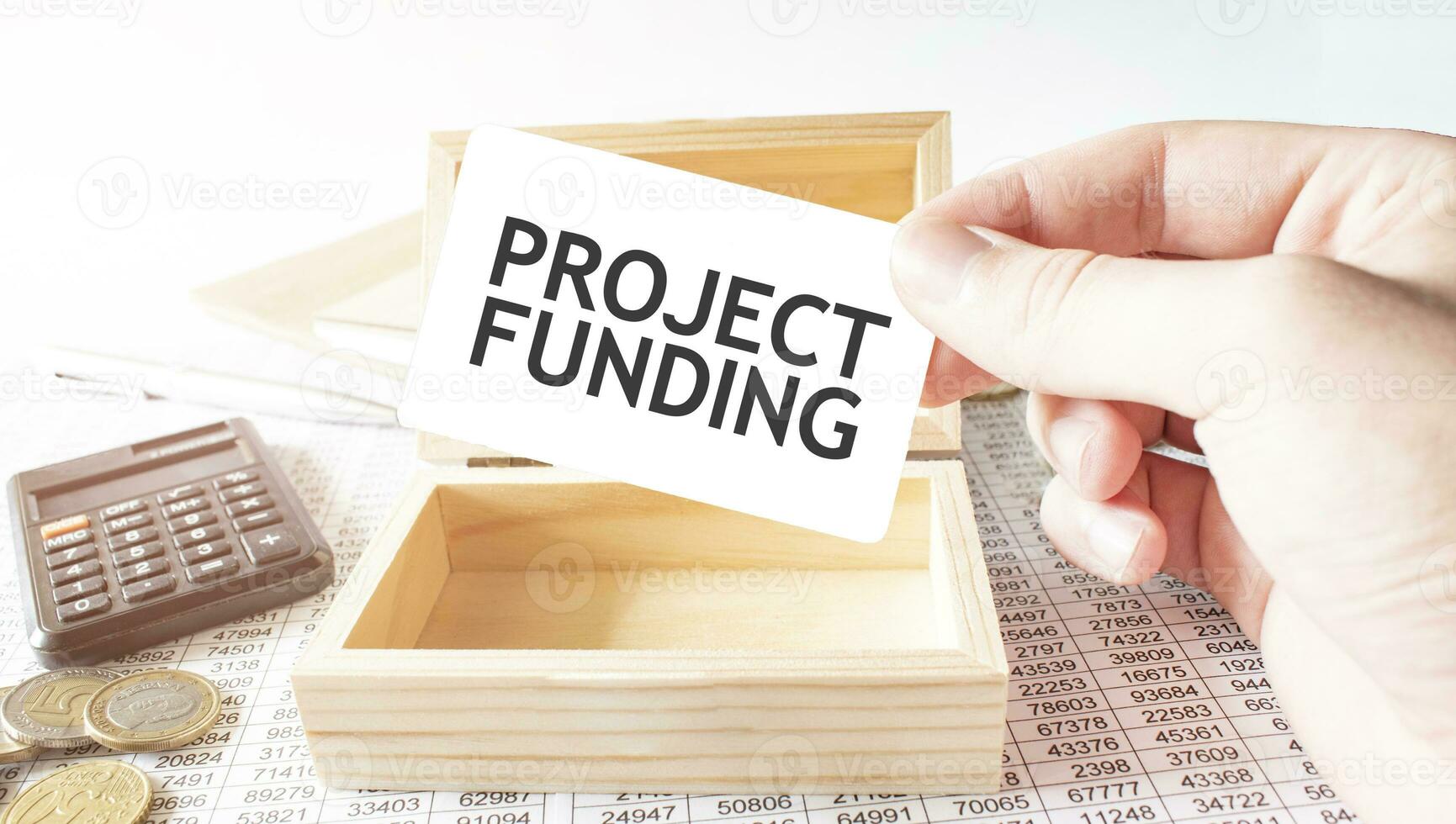 Businessman hold white card with text PROJECT FUNDING Calculator, wood box, money and financial documents photo