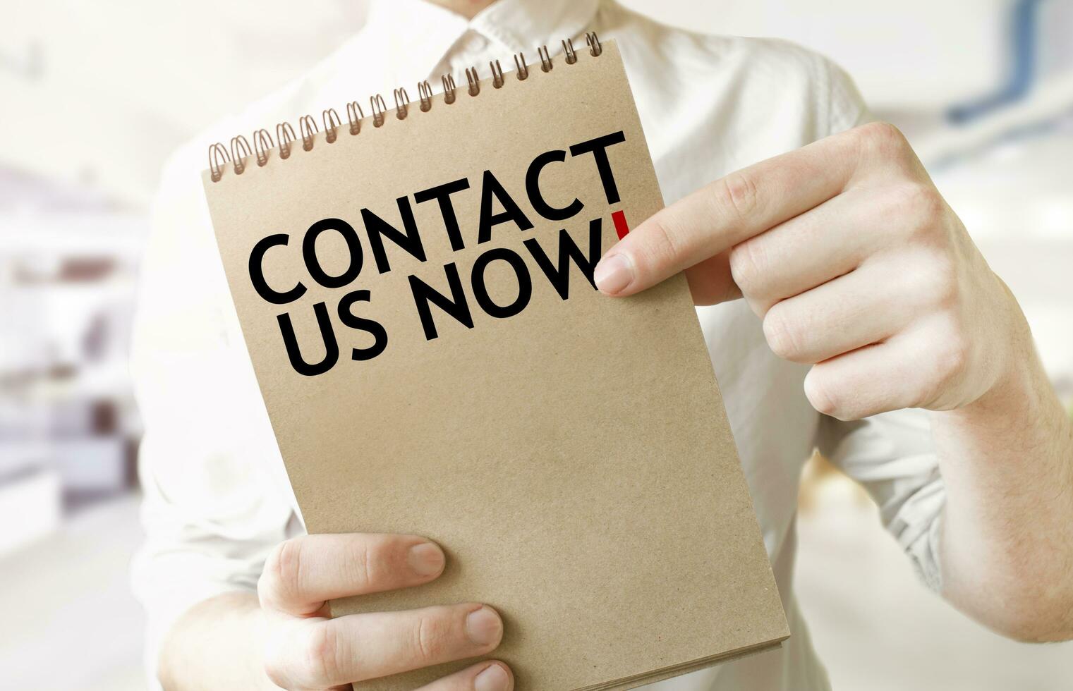 Text CONTACT US NOW on brown paper notepad in businessman hands in office. Business concept photo