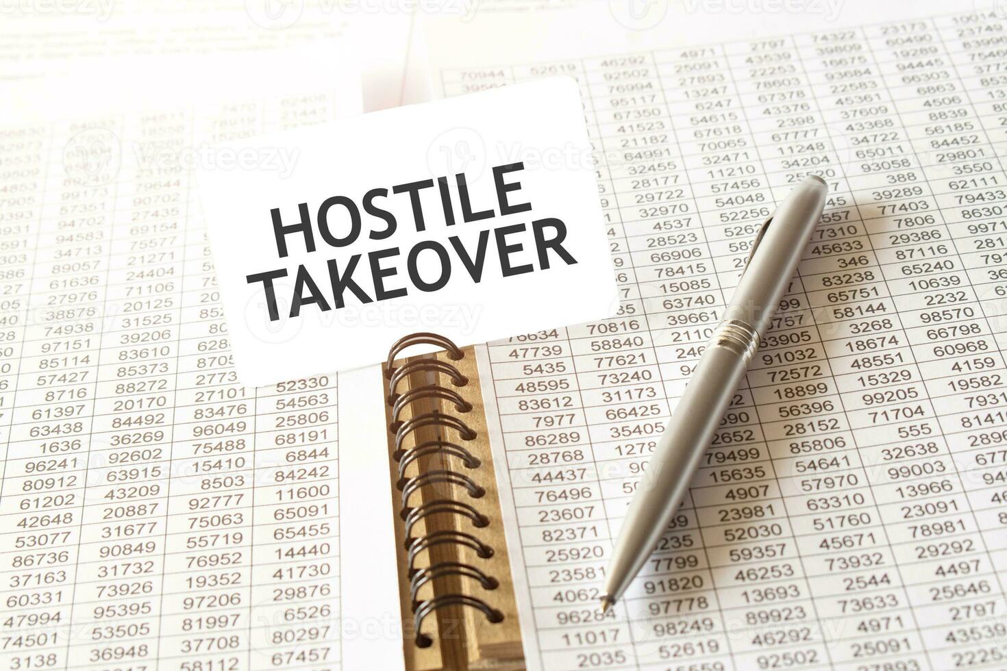 Text HOSTILE TAKEOVER on paper card, pen, financial documentation on table photo