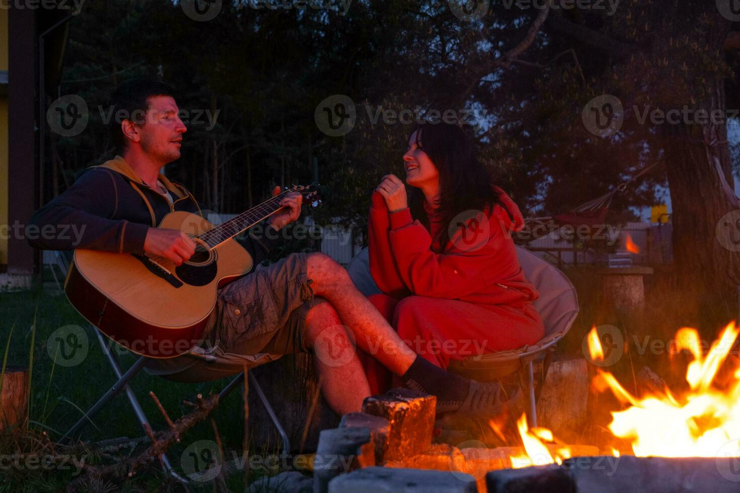 A man plays the guitar, a woman listens and sings along. A couple in love is sitting by the outdoor campfire in the courtyard of the house on camping chairs, a romantic evening photo