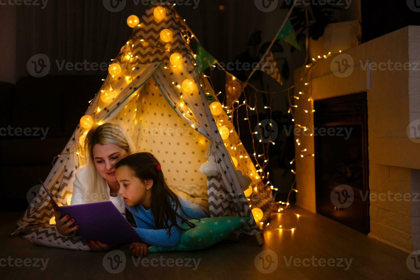 Mother and daughter are sitting in a teepee tent, reading stories with the flashlight. Happy family. photo