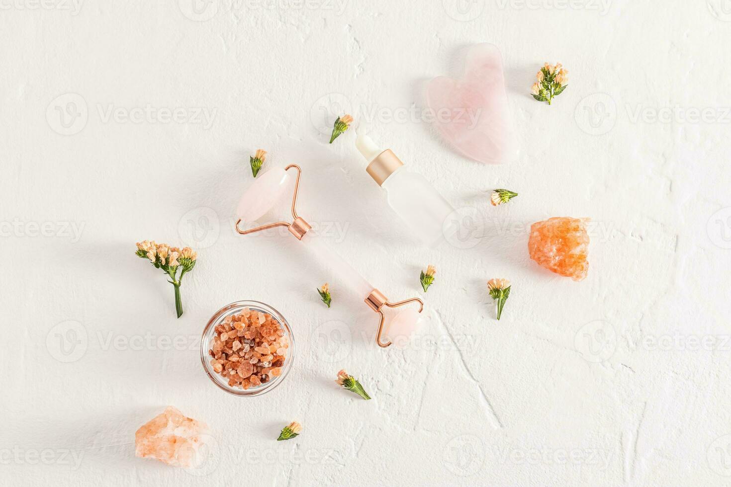 A set of cosmetics and a quartz roller massager with gua sha on a white textured background among flowers and lumps of bath salt. Top view. Flat lay. photo