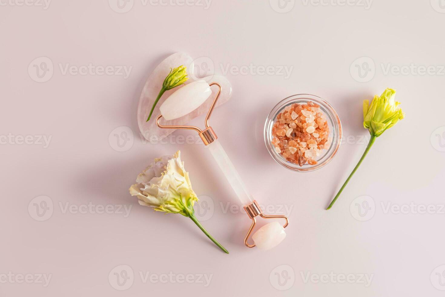 Cosmetic massager made of quartz, gua sha scraper, bath salt in a bowl on a pink background with astromeria flowers. Top view. Flat layout. photo