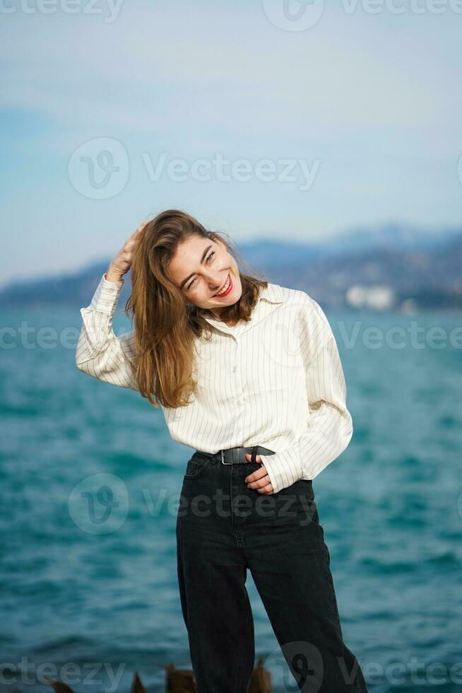 Beautiful young girl posing by sea. Girl smiling mischievously and looking squinted straight at camera. Girl wearing casual clothes. Walking by sea on sunny day. Happy young woman on holiday. photo