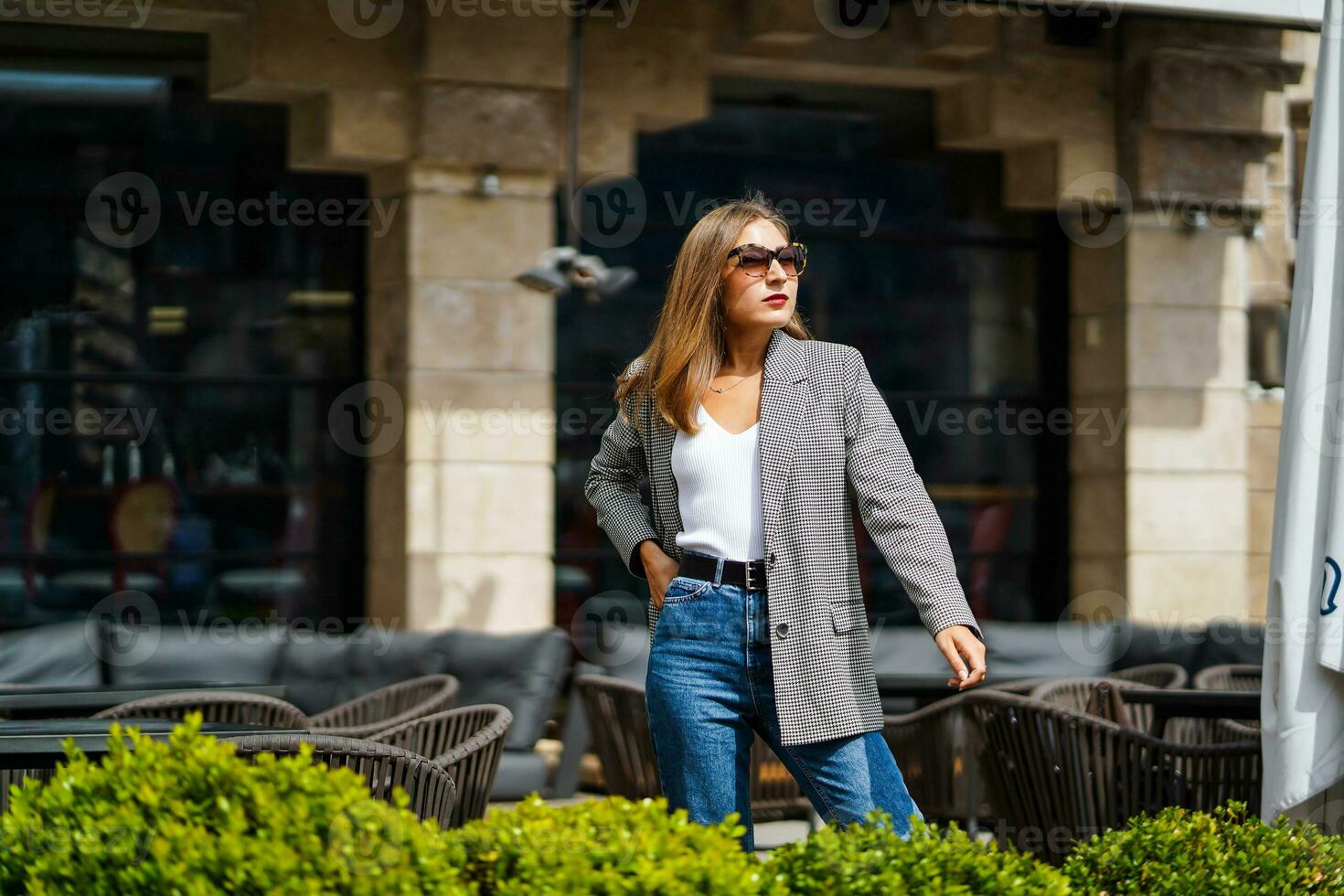 niña en pie cerca calle ciudad cafetería, calle sillas. maravilloso joven mujer en soleado homosexual estilo de vida concepto. elegante Moda negocio mujer con gris chaqueta, vaqueros. europeo café interior en antecedentes foto