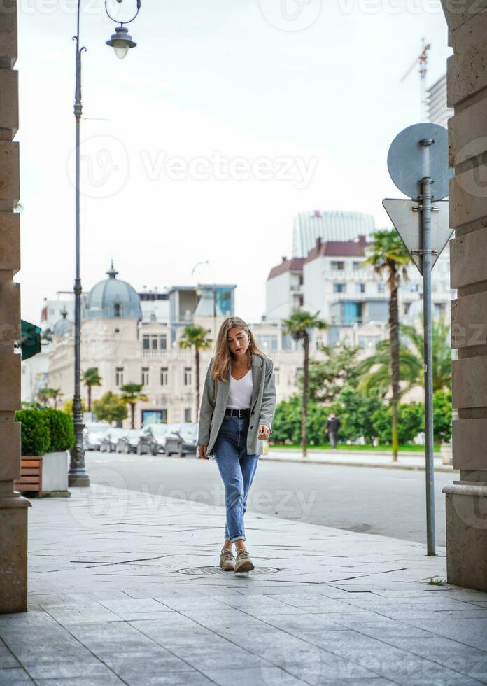 ciudad caminar. joven mujer caminando en acera en ciudad. niña vistiendo chaqueta y pantalones en arquitectónico edificios antecedentes. antiguo hermosa batumi ciudad en Georgia. foto