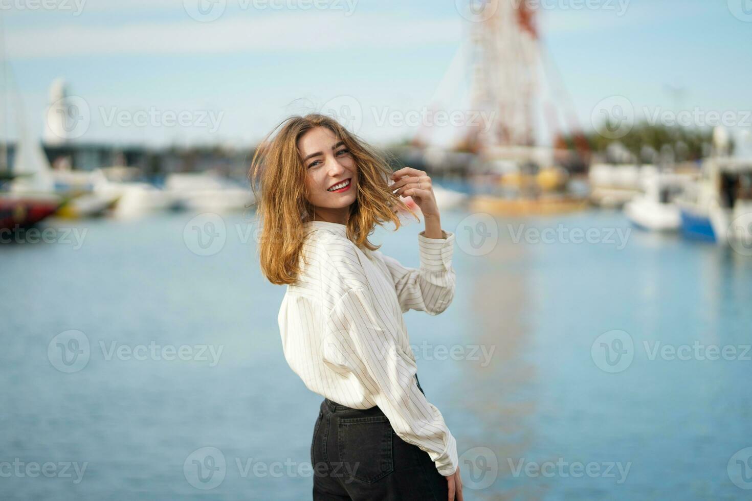 View from back. Young woman turned around and looking straight at camera. Cheerful pretty girl posing against seaport. Girl wearing casual clothes ang smilling. Walking by sea on sunny day. photo