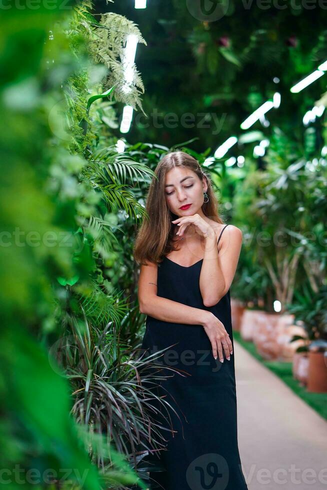 Thinking beautiful woman standing in thicket of tropical plants. Young woman wearing black dress on thin straps stands among green plants inside greenhouse. Girl With Her Eyes Down Thinking. photo