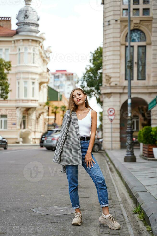 Girl threw jacket on her shoulder. Beautiful young woman wearing casual clothes standing on sidewalk. Historic building facade on background. City recreation. Person resting experiencing calmness photo