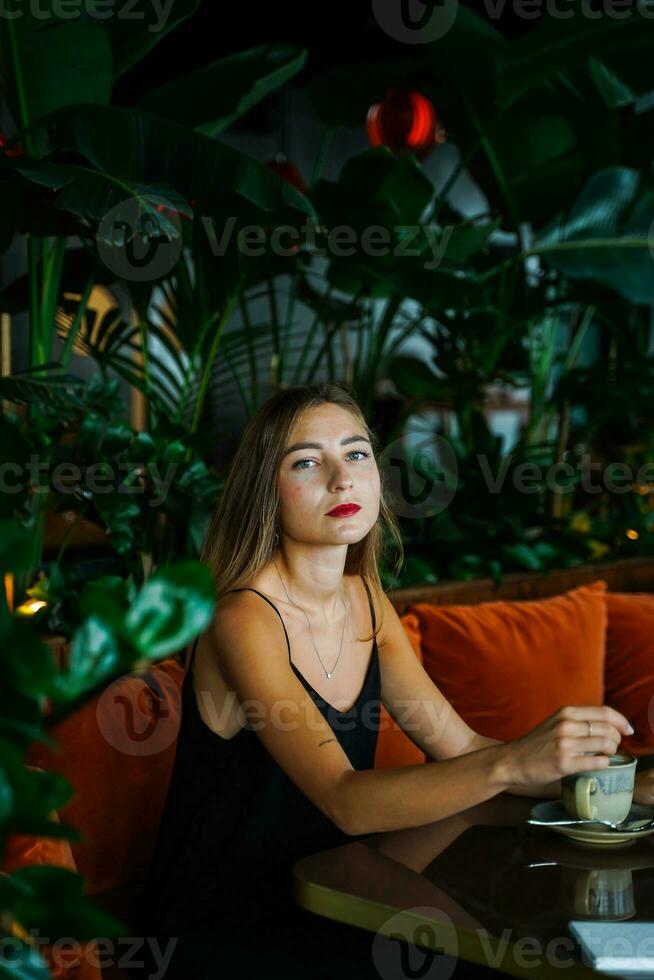 Young woman wearing black dress on thin straps siting on soft sofa at table in cafe among green plants inside arrangement. Thought Girl looking directly at camera. Hot drink cup on table. High quality photo
