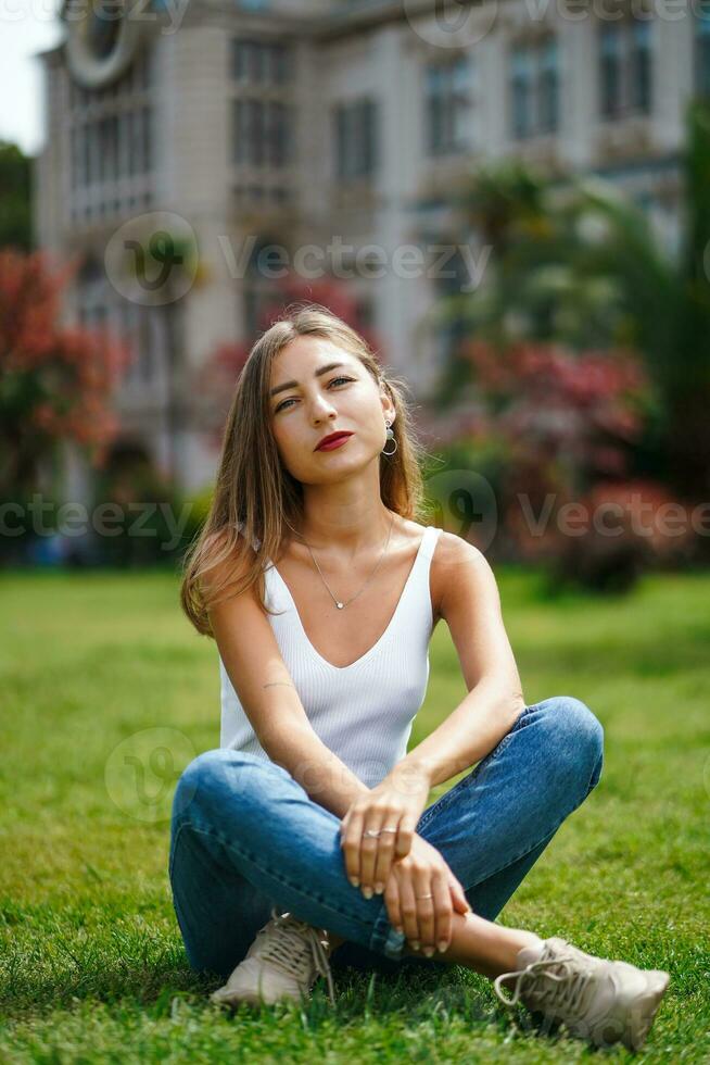 Beautiful young woman wearing tank top and jeans sitting on grass lawn in front of building. Young woman posing sitting on green grass. City recreation. Carefree person resting experiencing calmness photo
