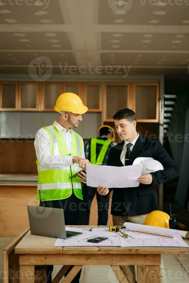 Contractor and architect looking at plan, tablet, laptop and talking about working on big project. Building in construction interior. photo